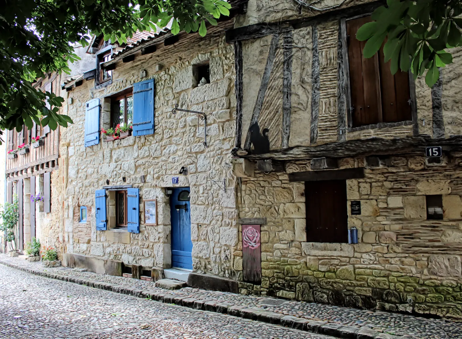 Authentic medieval stone house in Bergerac, France, featuring bright blue shutters and timber framing on a cobblestone street. Small black cat silhouette and a painted pink rose detail on the facade."