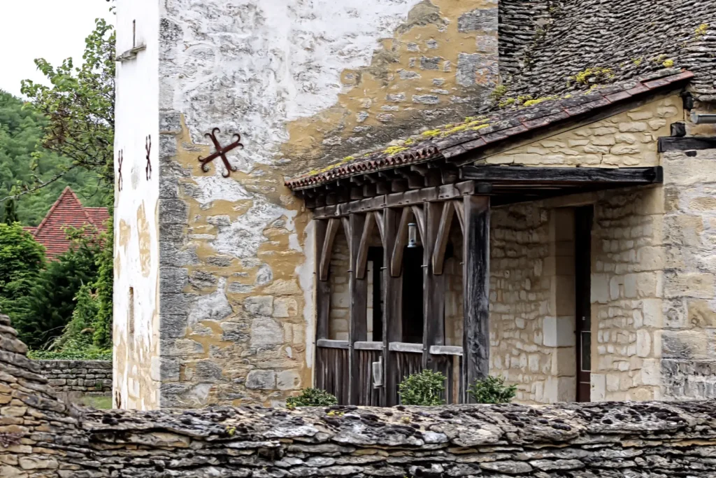 Close-up of a historic stone house in the Dordogne region of France, featuring a traditional lauze stone roof, a weathered wooden porch, and a decorative wrought iron wall anchor on a textured limestone facade."